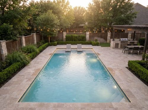 Rendering of large rectangular pool with a wide tanning ledge and expansive travertine deck on three sides, an outdoor kitchen and dining area, string lights and pool lighting at dusk, light blue water, deck jets arcing into the pool