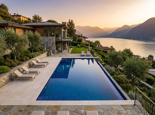 Rendering of rectangular infinity-edge pool on a hillside with the far edge vanishing into a lake and mountain view, glass tile finish with vivid blue water, limestone deck, modern house, golden hour