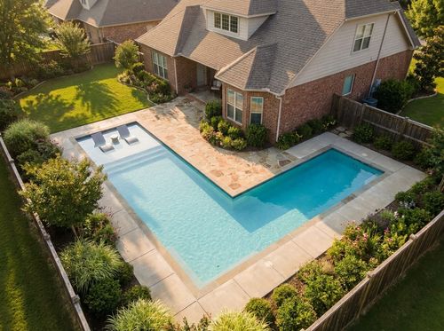 Rendering of L-shaped in-ground pool wrapping around a stone patio, light blue quartz water, broom-finished concrete deck, shallow leg with a tanning ledge, deeper long leg, traditional two-story house