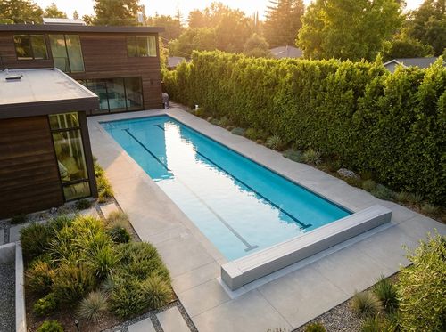 Rendering of 10 by 50 foot lap pool with lane lines, light blue quartz water, minimalist concrete deck, an automatic cover housing at one end, modern house, tall privacy hedge