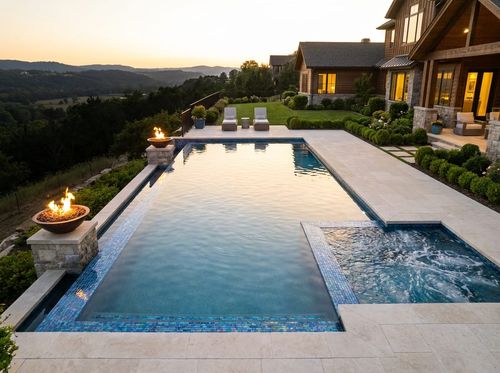 Rendering of luxury rectangular pool with an infinity edge overlooking a valley view, iridescent glass tile with vivid blue water, attached spa, two fire bowls on stone pedestals, limestone deck, modern estate house at dusk