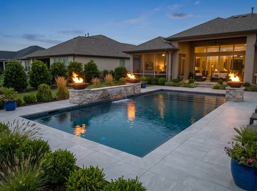 Rendering of geometric pool at dusk with two gas fire bowls burning on stone pedestals at the far corners reflected in dark blue water, porcelain paver deck, sheer descent on a low wall, modern house lit from within